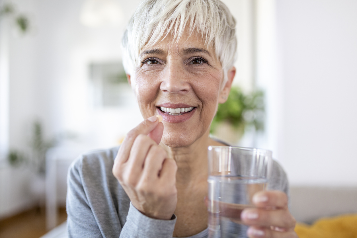 Smiling happy healthy middle aged 50s woman holding glass of water taking dietary supplement vitamin pill. Old women multivitamins antioxidants for anti age beauty.